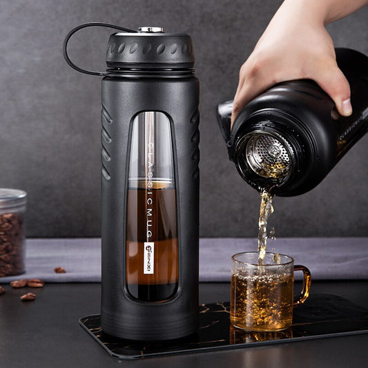 Eine Hand giesst Tee aus einer doppelwandigen, isolierten Glasflasche mit schwarzem Teesieb in eine klare Teetasse. Die Flasche hat eine schwarze Schutzhuelle mit Griff it. Im Hintergrund ist eine dunkle Arbeitsplatte und Kaffeebohnen zu sehen.