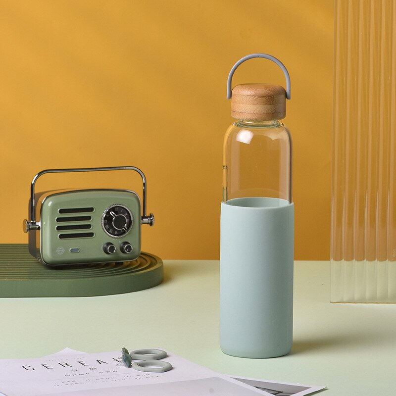 Eine Glas-Trinkflasche mit Holzdeckel und mintgruenem Silikonueberzug, daneben ein gruenes Radio, vor einem gelben Hintergrund.