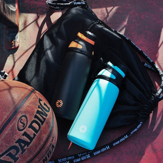 Das Bild zeigt zwei Thermosflaschen, eine in Schwarz und eine in Blau, neben einem Basketball und einer schwarzen Sporttasche auf einem roten Boden.
