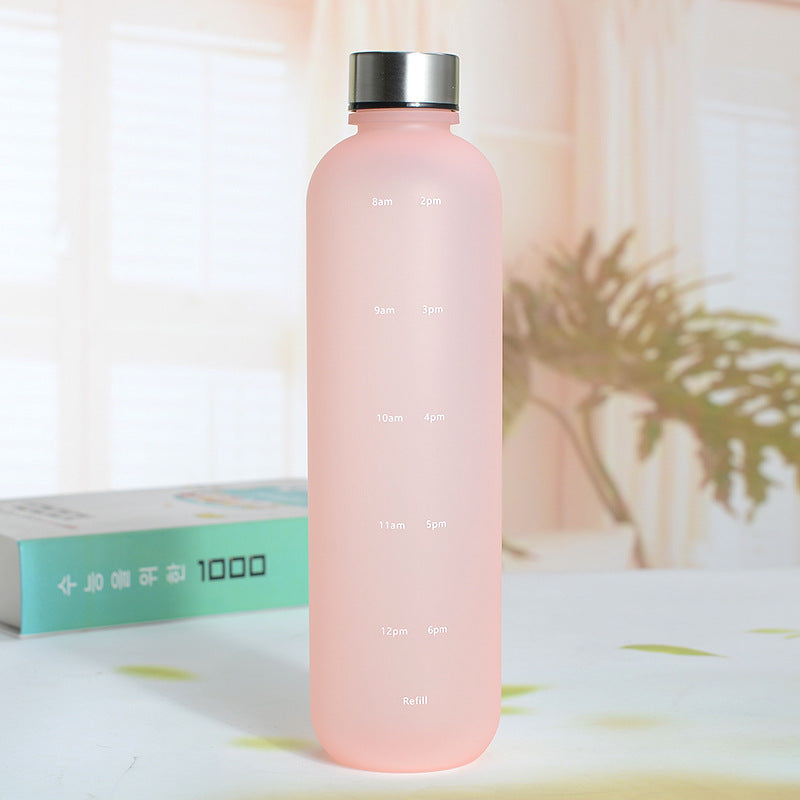 Eine Trinkflasche mit einer Farbe Pink und einem silbernen Deckel. Die Flasche hat weisse Zeitmarkierungen auf der Seite. Sie steht auf einer Oberflaeche mit einer unscharfen, hellen Umgebung und einer Pflanze im Hintergrund.