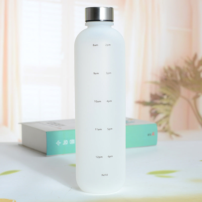 Eine Trinkflasche mit einer Farbe Weiss und einem silbernen Deckel. Die Flasche hat weisse Zeitmarkierungen auf der Seite. Sie steht auf einer Oberflaeche mit einer unscharfen, hellen Umgebung und einer Pflanze im Hintergrund.