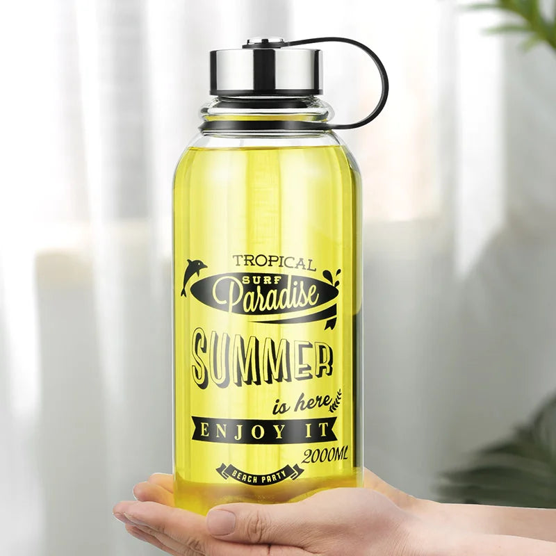 Eine handgehaltene gelbe Glaswasserflasche mit einem Fassungsvermoegen von 2000 ml. Auf der Flasche ist ein sommerliches Motiv mit der Aufschrift "Tropical Surf Paradise" und "SUMMER is here ENJOY IT" zu sehen.