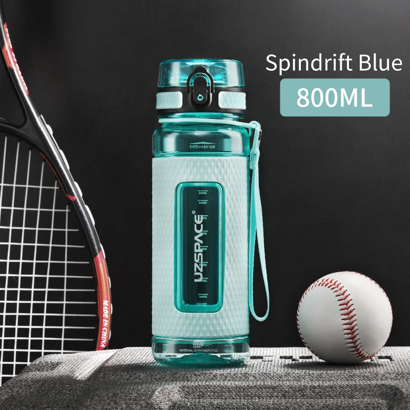 Eine blaue Sport-Trinkflasche mit einem Fassungsvermoegen von 0.8 Litern, gekennzeichnet durch das Logo "UZSPACE" im klaren Sichtfenster, platziert zwischen einem Tennisracket und einem Baseball auf einer grauen strukturierten Oberflaeche vor einem schwarzen Hintergrund.