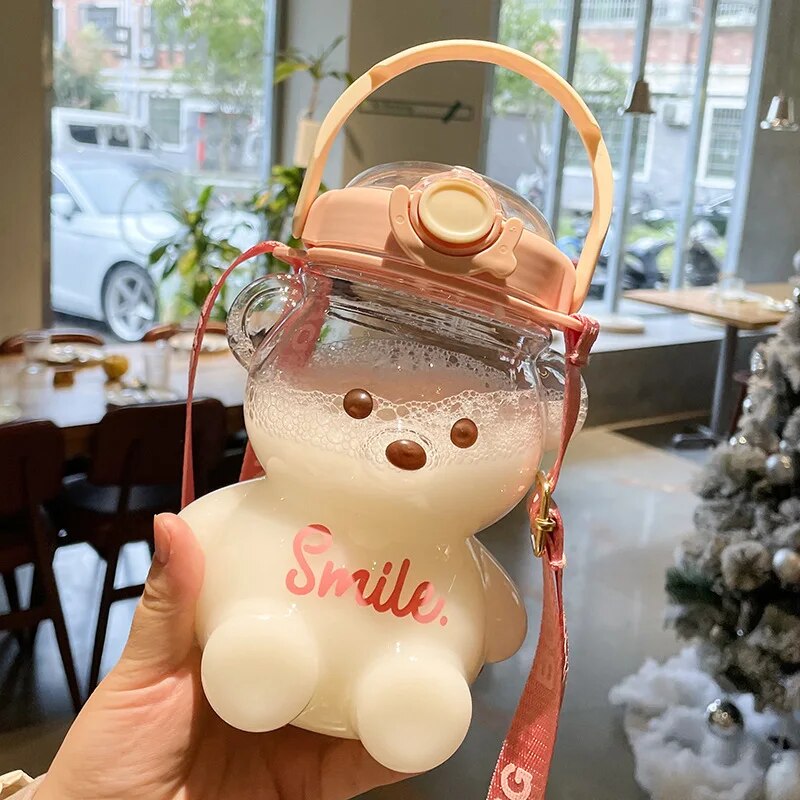 Hand haelt eine durchsichtige, weiße Trinkflasche in Teddybaerform mit einem rosa Deckel und einem 'Smile.'-Schriftzug."