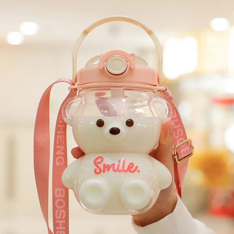 Hand haelt eine transparente, weiße Trinkflasche für Kinder in Teddybaerform mit rosa Deckel und Schriftzug 'Smile' an einem rosa Umhaengeband.