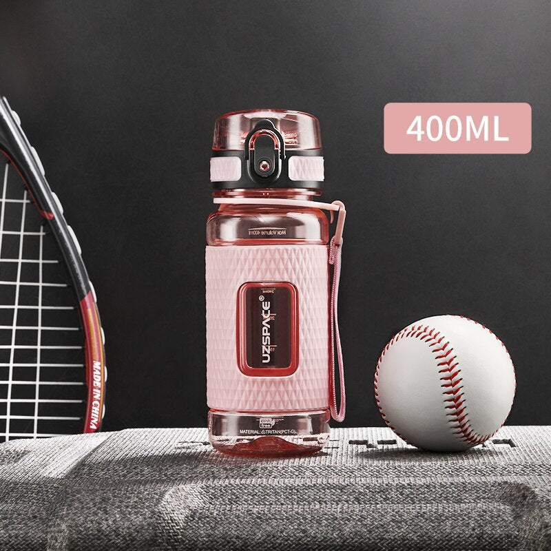 Eine pinke Sport-Trinkflasche mit einem Fassungsvermoegen von 0.4 Litern, gekennzeichnet durch das Logo "UZSPACE" im klaren Sichtfenster, platziert zwischen einem Tennisracket und einem Baseball auf einer grauen strukturierten Oberflaeche vor einem schwarzen Hintergrund.
