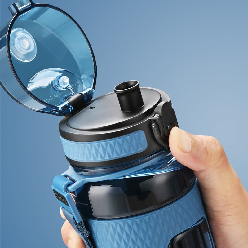 Eine Hand haelt eine blaue Sport-Trinkflasche mit Sicherheitsverschluss und transparentem Klappdeckel vor einem blauen Hintergrund.