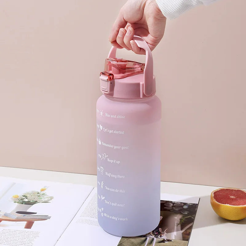 Eine Hand haelt eine grosse, rosafarbene Sporttrinkflasche mit motivierenden Zeitmarkierungen auf einem offenen Buch und Tisch neben einer halbierten Grapefruit.