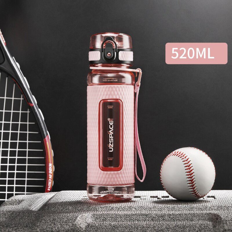 Eine pinke Sport-Trinkflasche mit einem Fassungsvermoegen von 0.52 Litern, gekennzeichnet durch das Logo "UZSPACE" im klaren Sichtfenster, platziert zwischen einem Tennisracket und einem Baseball auf einer grauen strukturierten Oberflaeche vor einem schwarzen Hintergrund.