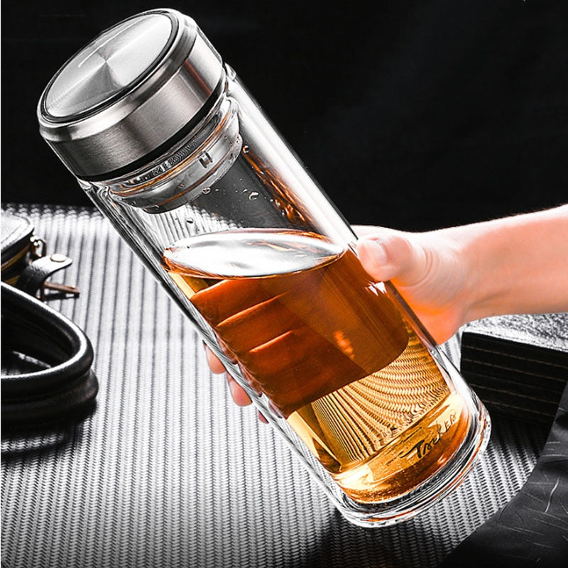 Eine Hand haelt eine transparente Glasflasche mit Tee. Der Tee hat eine goldene Farbe. Die Flasche hat einen metallischen Schraubdeckel und ein integriertes Teesieb. Sie steht vor einem stilvollen, dunklen Hintergrund mit strukturierter Oberflaeche.