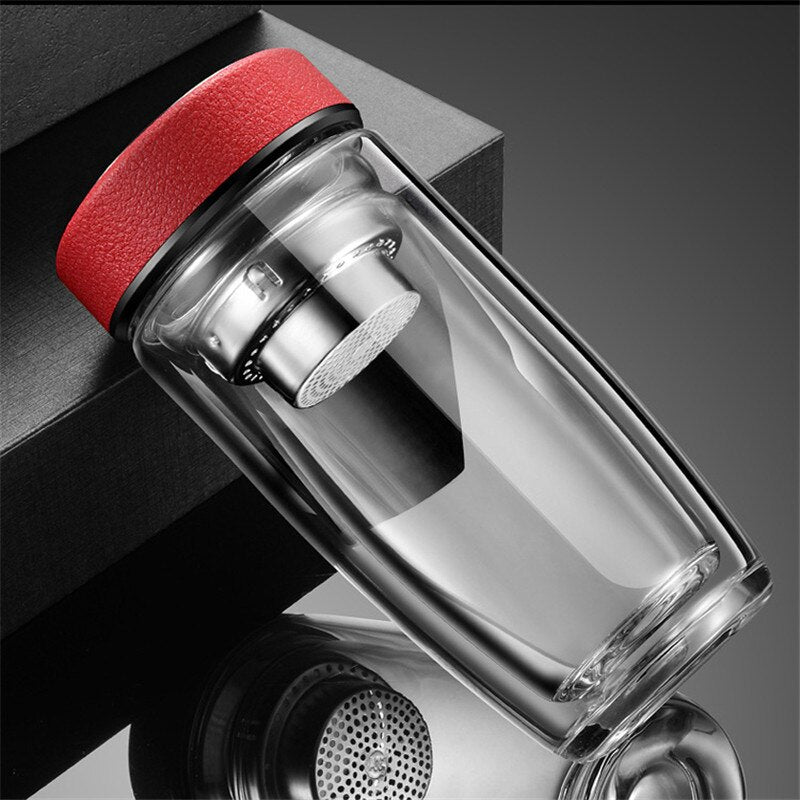 Eine Glasflasche mit integriertem Teesieb und einem strukturierten rotem Deckel, praesentiert auf einer reflektierenden Oberflaeche neben einer schwarzen Box.
