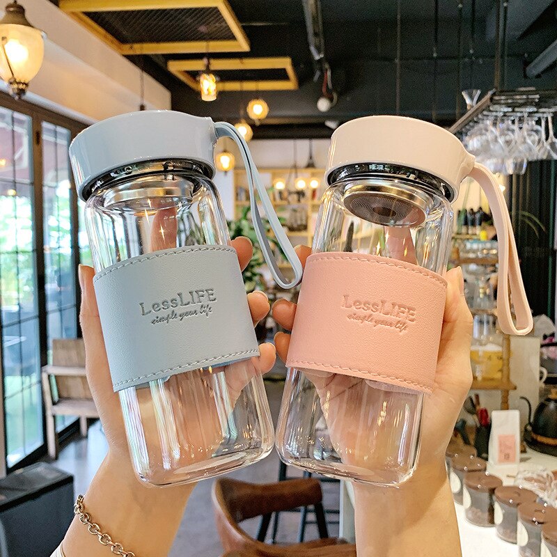 Zwei Haende halten moderne Glas-Trinkflaschen mit Teesieben, die jeweils mit einer hellblauen und einer rosa Schutzhuelle versehen sind. Im Hintergrund ist eine Cafe-Umgebung mit Beleuchtung und verschwommenen Einrichtungsgegenstaenden zu erkennen.