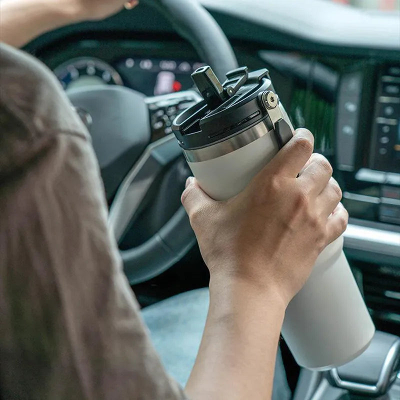Person im Fahrzeug haelt eine weiße Thermos-Trinkflasche mit Trinkhalm und Edelstahldeckel waehrend der Fahrt.