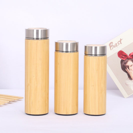 Drei Thermosflaschen mit Bambusummantelung und Edelstahldeckeln in unterschiedlichen Groeßen. Im Hintergrund ist eine Postkarte mit einer abgebildeten Person zu sehen.