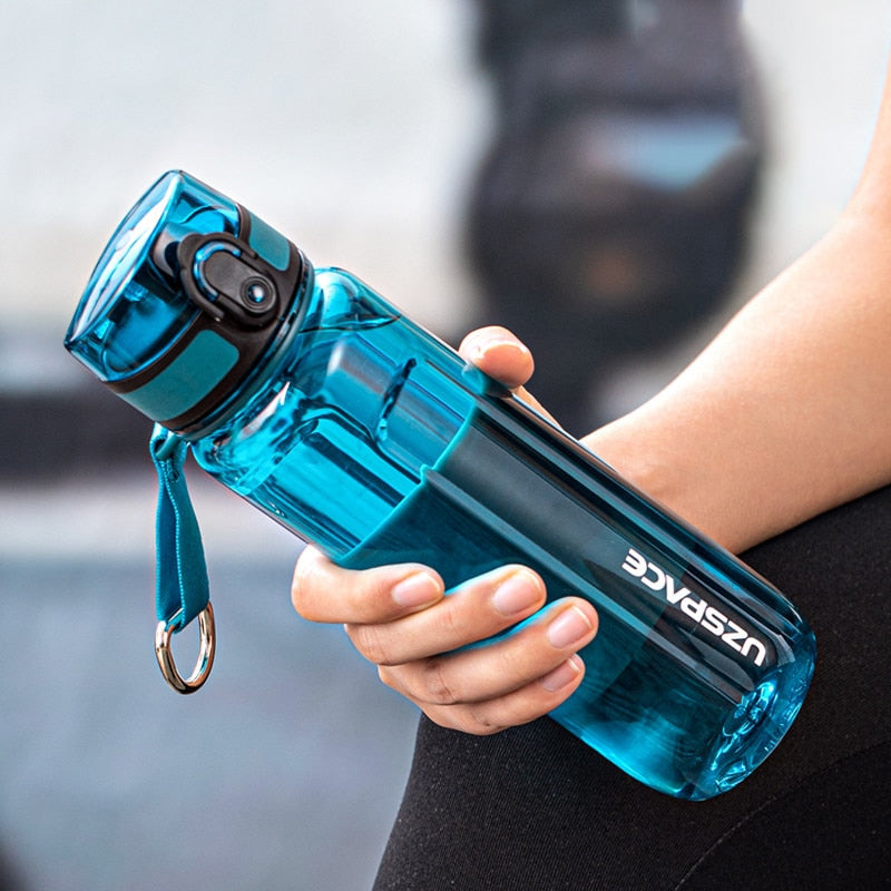 Eine Hand haelt eine transparente tüerkies Sporttrinkflasche mit einem Trageband und einem Klappdeckel. Auf der Seite der Flasche ist das umgedrehte Logo "UZSPACE" sichtbar.