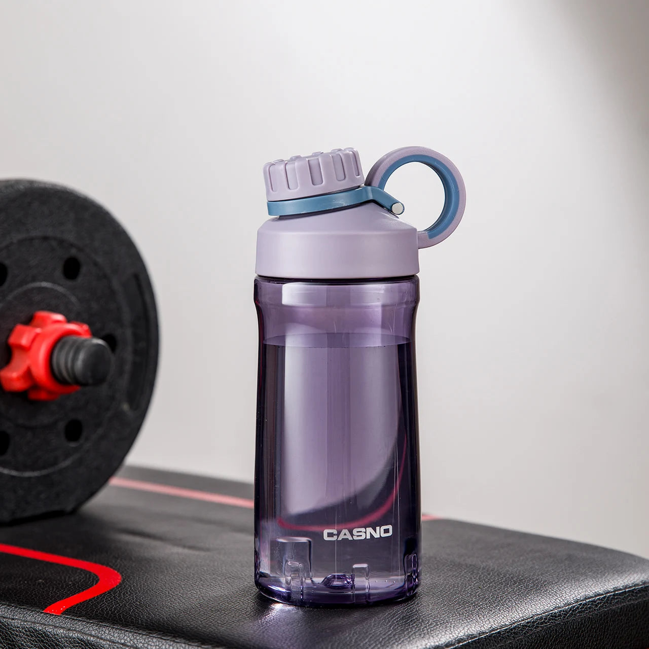 Eine violette, transparente Sporttrinkflasche auf einer Fitnessbank. Die Flasche hat einen schwarzen Schraubverschluss mit einem Henkel und ist mit dem Wort "SPORT" sowie der Marke "CASNO" beschriftet. Im Hintergrund ist eine Hantel mit roten Details zu sehen.