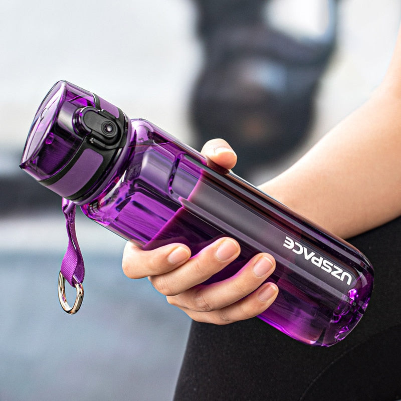 Eine Hand haelt eine transparente violette Sporttrinkflasche mit einem Trageband und einem Klappdeckel. Auf der Seite der Flasche ist das umgedrehte Logo "UZSPACE" sichtbar.
