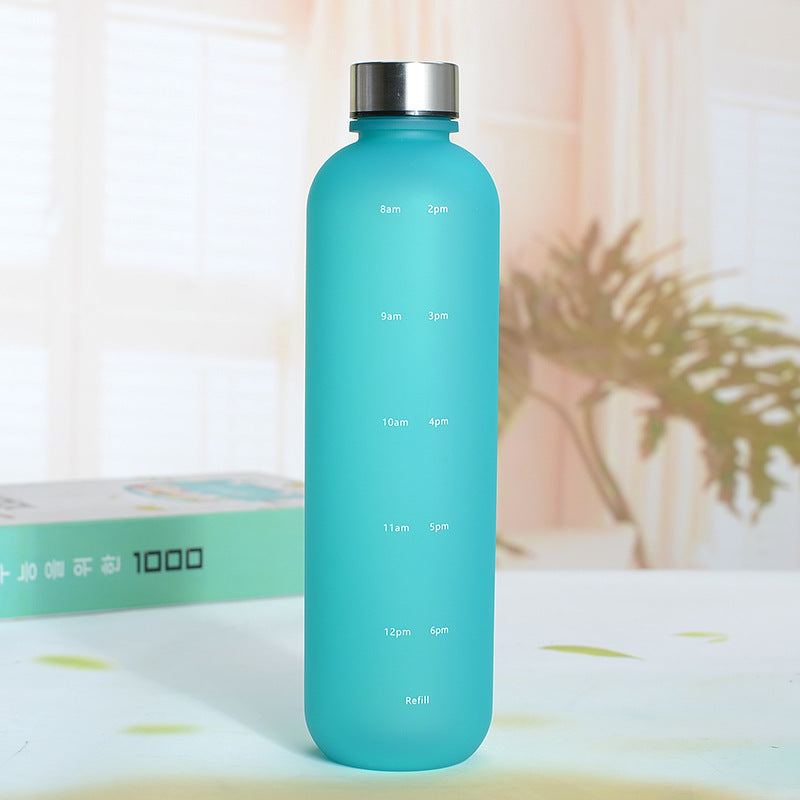Eine Trinkflasche mit einer Farbe Blau und einem silbernen Deckel. Die Flasche hat weisse Zeitmarkierungen auf der Seite. Sie steht auf einer Oberflaeche mit einer unscharfen, hellen Umgebung und einer Pflanze im Hintergrund.
