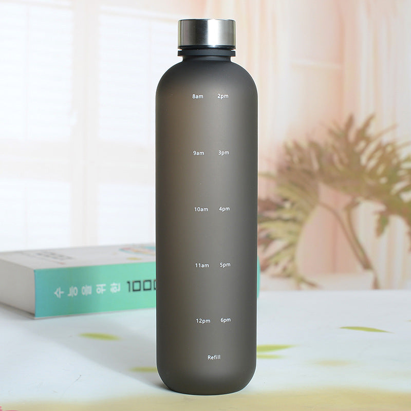 Eine Trinkflasche mit einer Farbe Schwarz und einem silbernen Deckel. Die Flasche hat weisse Zeitmarkierungen auf der Seite. Sie steht auf einer Oberflaeche mit einer unscharfen, hellen Umgebung und einer Pflanze im Hintergrund.
